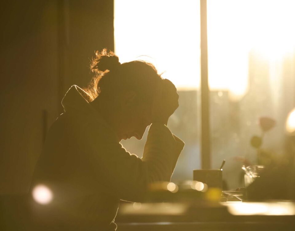 Side view of anonymous woman silhouette working at table with supplies and cup of hot drink while leaning on hands in bright back lit of sunset light at home. She is tired